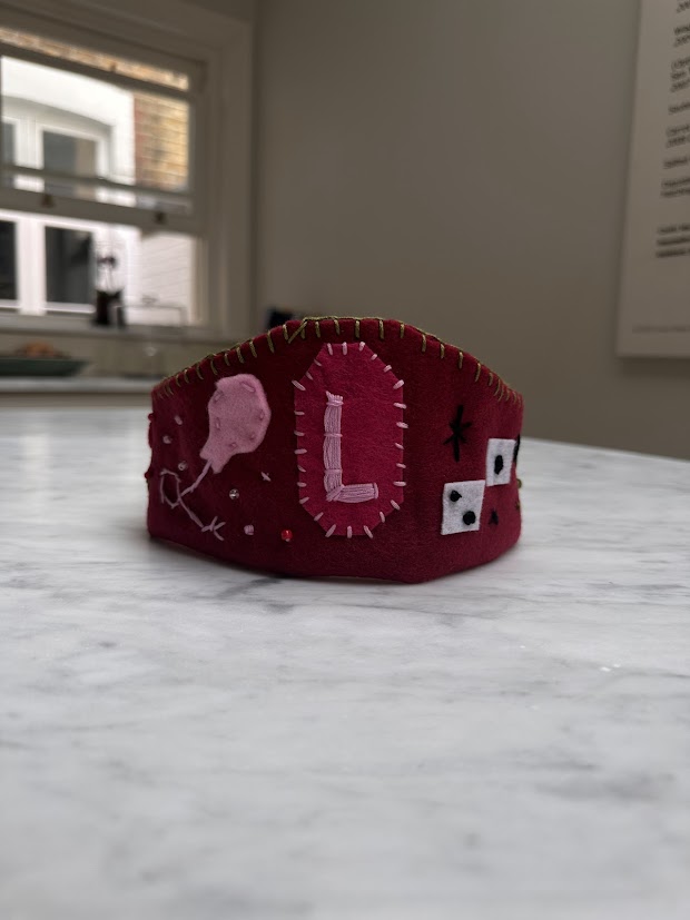 Deep red felt diadem crown with a pink L, balloon and dice embroidery